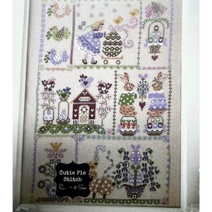 May include: A framed cross-stitch embroidery featuring a colorful Easter theme with bunnies, chicks, eggs, and flowers. The embroidery is titled "Cutie Pie Stitch" and signed "C & Sue".
