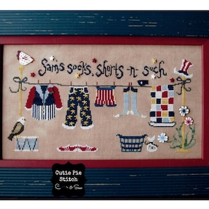 May include: A framed cross-stitch design featuring a clothesline with various items hanging on it, including socks, shorts, and a towel. The text "Sam's socks, shorts 'n such" is stitched above the clothesline. The design is framed in a dark blue frame with a red mat.