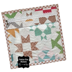 May include: A quilted zippered pouch with a patchwork design in shades of brown, green, and orange. The pouch features a white lace trim and a brown and white gingham border. The text "Cutie Pie Stitch & Sew" is visible.