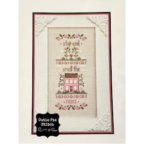May include: A framed cross-stitch design with a pink house and flowers. The text reads "stop and smell the roses".
