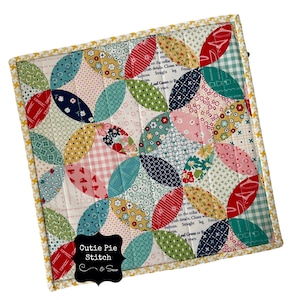 May include: A colorful patchwork quilt block with a "Cutie Pie Stitch" label. The block features a repeating pattern of curved shapes in shades of red, pink, blue, green, yellow, and white.