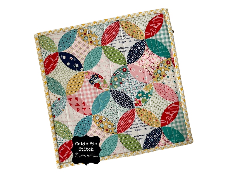 May include: A colorful patchwork quilt block with a "Cutie Pie Stitch" label. The block features a repeating pattern of curved shapes in shades of red, pink, blue, green, yellow, and white.