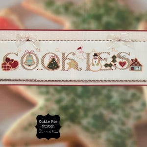 Peut inclure: Un sampler de point de croix blanc avec le mot "COOKIES" en lettres brunes. Chaque lettre est décorée d'une petite image sur le thème de Noël, y compris une cloche, un sapin de Noël, un Père Noël, un bonhomme de neige et une maison en pain d'épices.