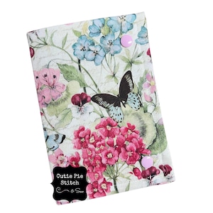May include: A floral-patterned notebook cover featuring pink and blue flowers, butterflies, and green leaves. The cover has two pink push pins and the text "Cutie Pie Stitch & Sew" in a black banner.