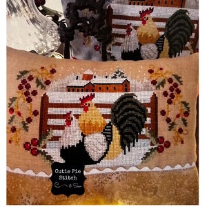 May include: A cross-stitched pillow featuring a winter scene with roosters, a snow-covered barn, and a wooden fence. The pillow has a tan background with a scalloped edge and the text "Cutie Pie Stitch & Sew".