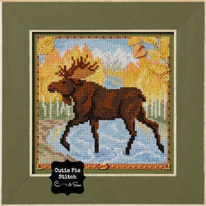 May include: A cross-stitch framed artwork featuring a brown moose standing in a blue and white stream. The background is a yellow and orange autumn forest with a single brown leaf in the upper right corner. The artwork is titled "Cutie Pie Stitch & Sew".