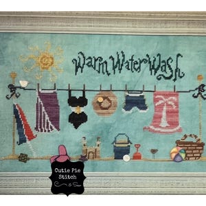 May include: A cross-stitch embroidery design featuring a clothesline with various beach-themed items hanging on it, including a swimsuit, a towel, and a hat. The text "Warm Water Wash" is stitched above the clothesline. The design is framed in a light wood frame.