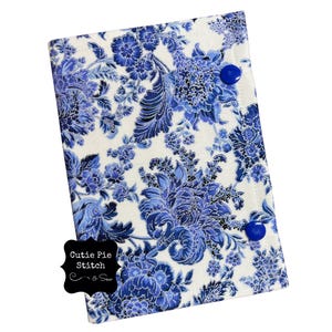 May include: A fabric journal cover with a blue and white floral pattern. The cover is secured with two blue buttons. The words "Cutie Pie Stitch & Sew" are printed on a black label in the lower left corner.