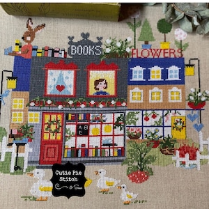 May include: A cross-stitch image of a charming bookstore and florist. The buildings are detailed with windows, doors, and decorative elements. A rabbit sits atop the bookstore, and ducks are in the foreground. The signs read "BOOKS" and "FLOWERS".