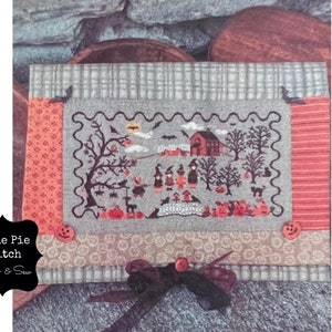 May include: A cross-stitch pattern featuring a spooky Halloween scene with a house, trees, and figures in black and orange on a gray background. The pattern is framed with a scalloped border and surrounded by orange and gray fabric.