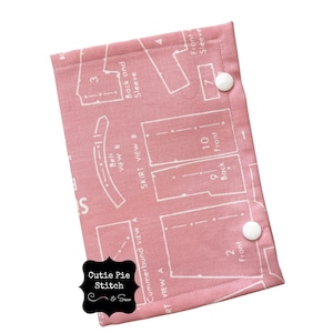 May include: Pink fabric with white sewing pattern markings. The fabric has two white snap buttons. The fabric has the text "Cutie Pie Stitch & Sew" printed on it.