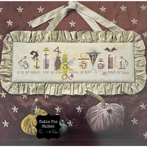 May include: A cross-stitch sampler with a Halloween theme. The sampler features a variety of Halloween-themed images, including a witch, a cat, a bat, a cauldron, and a pumpkin. The sampler is framed in a white frame with a gold trim. The text on the sampler reads "SI 4 la EYE OF NEWT TOE OF FRO HOOL OF BAT MAIR OF DOG".