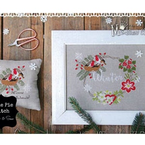May include: A white framed cross-stitch embroidery with a winter theme. The embroidery features two red robins perched on a branch with red berries, green leaves, and white flowers. The word "Winter" is stitched in white thread. The embroidery is on a beige linen fabric. The frame is on a wooden surface with a small pillow with a similar embroidery design.