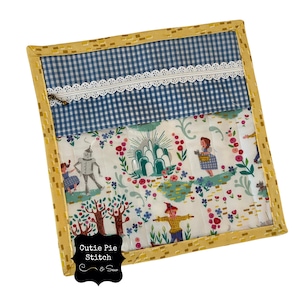 May include: A square, quilted fabric item with a yellow border and a blue and white gingham top section. The lower section features a colourful print with characters and scenes from The Wizard of Oz. The item has the text "Cutie Pie Stitch & Sew".