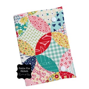 Puede incluir: Estuche de tela acolchada y colorida con un diseño de patchwork que presenta varios patrones y colores, incluyendo rojo, rosa, azul y amarillo. El estuche tiene dos cierres de botón blancos y una etiqueta negra con el texto "Cutie Pie Stitch & Sew".