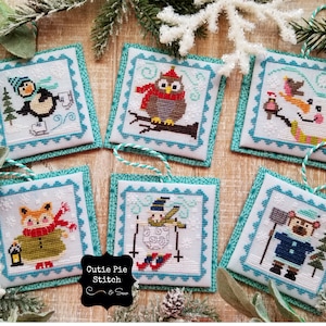 May include: Six handmade cross-stitch ornaments with winter themes. Each ornament features a different animal, including a penguin, owl, snowman, fox, and bear. The ornaments are framed with a teal blue border and have a white background with snowflakes.