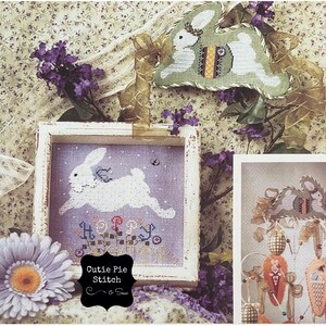 May include: A cross stitch pattern featuring a white bunny hopping with the word "Hoppy" in colorful letters. The pattern is framed in a white wooden frame and is surrounded by purple flowers. The text "Cutie Pie Stitch" is written on a black chalkboard.