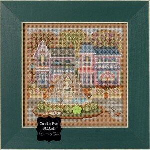 May include: A framed cross-stitch depicting a town square with buildings, a fountain, and trees. The buildings are in pastel colors, with the words "Millinery" and "Ice Cream" visible. The frame is dark green, and the artwork is signed "Cutie Pie Stitch & Sew."
