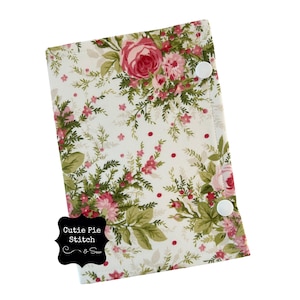 May include: A floral print case with a white background, featuring pink roses and green leaves. The case has two white snap closures. The text "Cutie Pie Stitch & Sew" is on a black label.