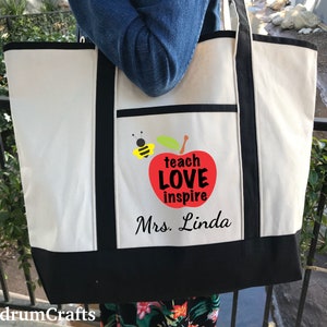 May include: A white canvas tote bag with black trim and a red apple graphic. The apple has the words "teach love inspire" on it and a bee is flying next to it. The text "Mrs. Linda" is printed below the apple.