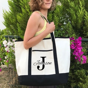 May include: A white canvas tote bag with black trim and a large black letter "J" with the name "Johnson" printed below. The bag is being held by a person wearing sunglasses.