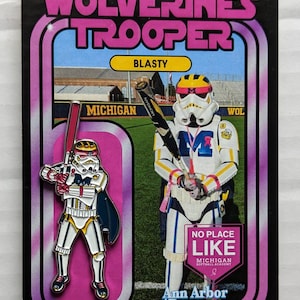 May include: A pink and black card with a white and yellow cartoon character holding a baseball bat. The character is wearing a helmet and a cape. The card says "Wolverine Trooper" and "Blast" in yellow letters. The card also says "No Place Like Michigan Softball Academy" in white letters on a pink background.