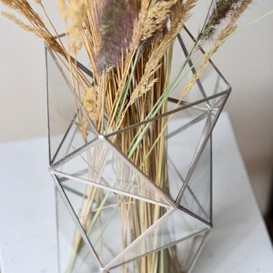 May include: A geometric glass vase with a silver frame holds a bouquet of dried grasses and wheat.