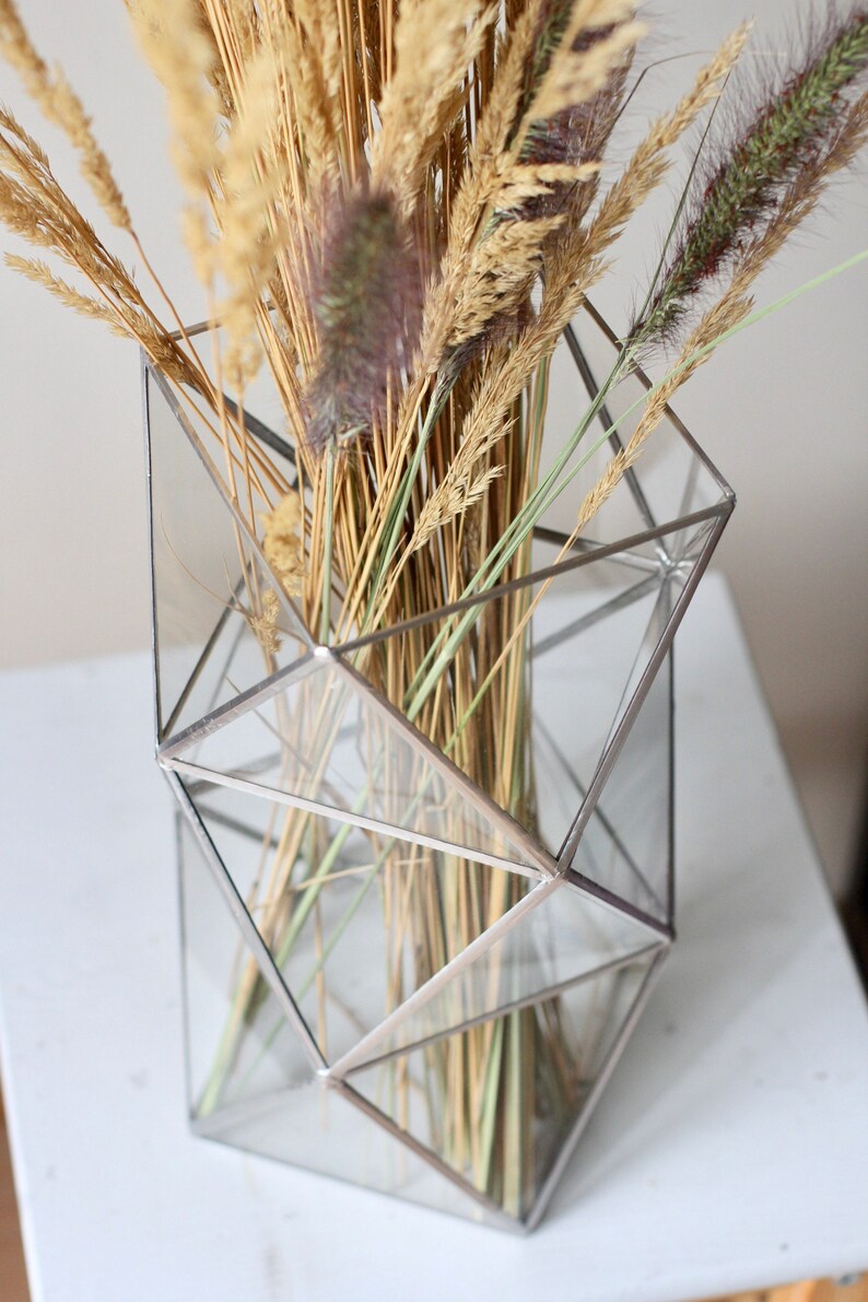 May include: A geometric glass vase with a silver frame holds a bouquet of dried grasses and wheat.