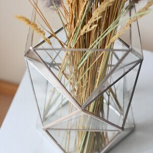 May include: A geometric glass vase with a silver frame holds a bouquet of dried grasses and wheat.