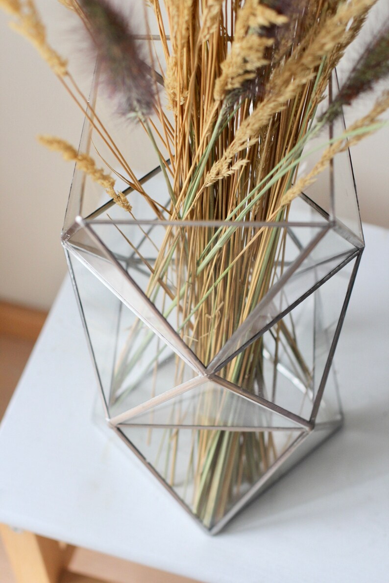 May include: A geometric glass vase with a silver frame holds a bouquet of dried grasses and wheat.