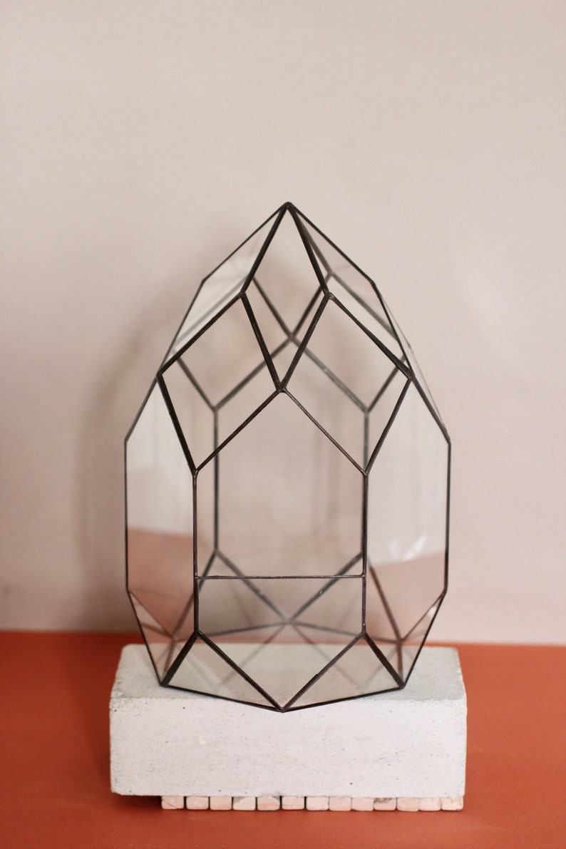 May include: Geometric glass terrarium with a black metal frame, resting on a white rectangular base. The clear glass panels create a multi-faceted shape, perfect for displaying small plants or decorative items. The terrarium is approximately 12 inches tall.