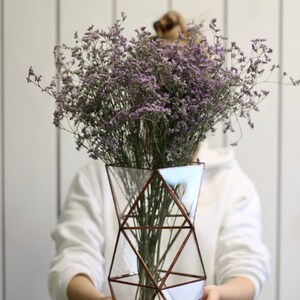 May include: A bouquet of purple flowers in a geometric glass vase with copper accents. The vase is held by a person wearing a white sweater.