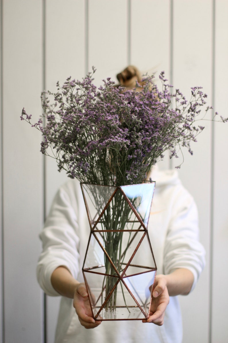 May include: A bouquet of purple flowers in a geometric glass vase with copper accents. The vase is held by a person wearing a white sweater.