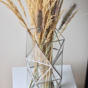 May include: A geometric glass vase with a silver frame holds a bouquet of dried grasses in shades of beige and brown.