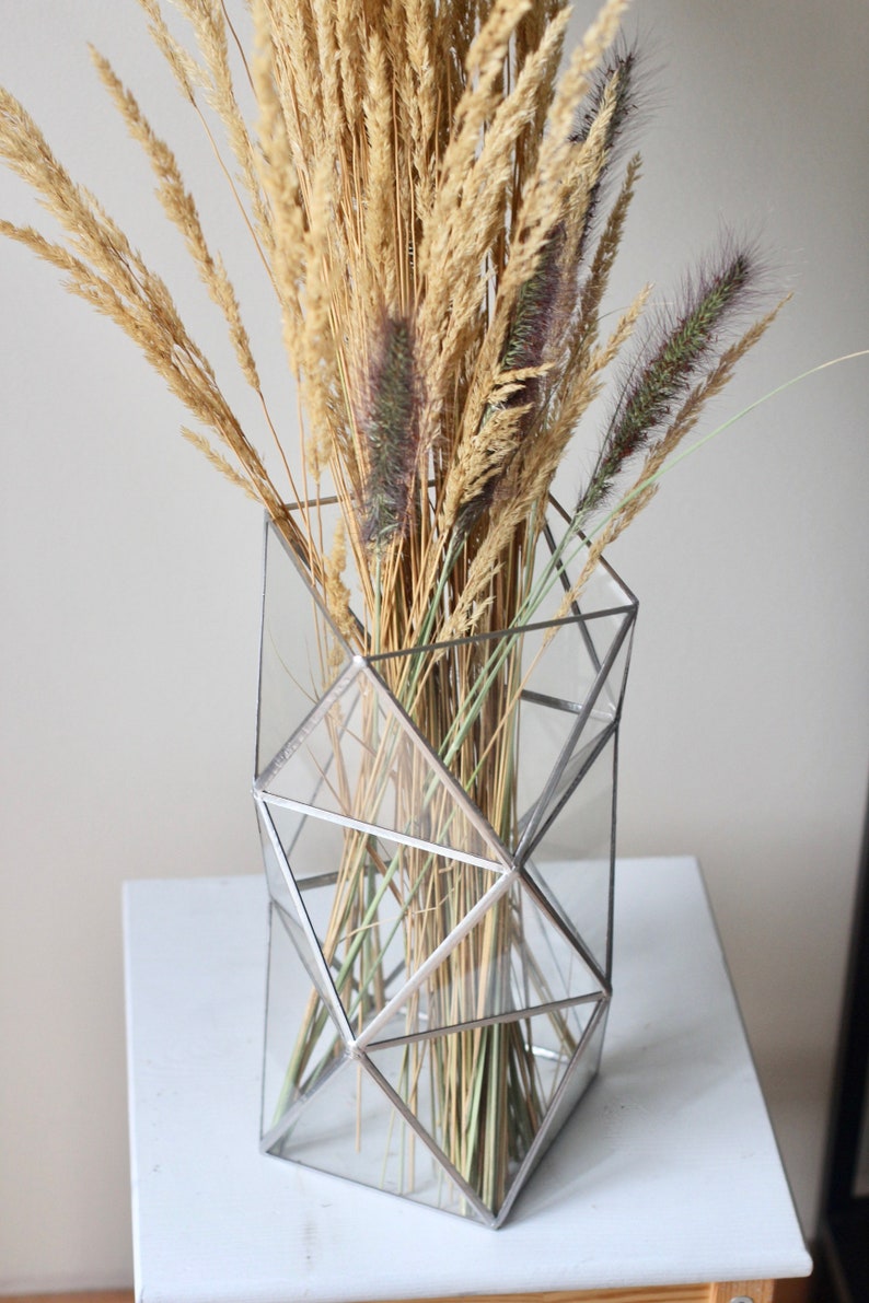 May include: A geometric glass vase with a silver frame holds a bouquet of dried grasses in shades of beige and brown.