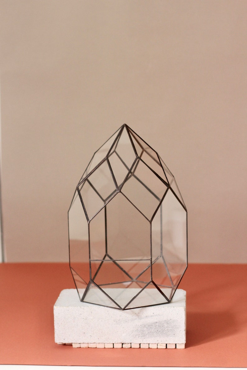 May include: Geometric glass terrarium with a black metal frame, resting on a white rectangular base. The terrarium has a pointed top and is designed with multiple clear glass panels.