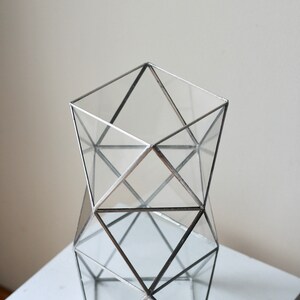 May include: A geometric glass vase with a silver metal frame. The vase is made of clear glass and has a square base. The frame is made of silver metal and has a geometric design.