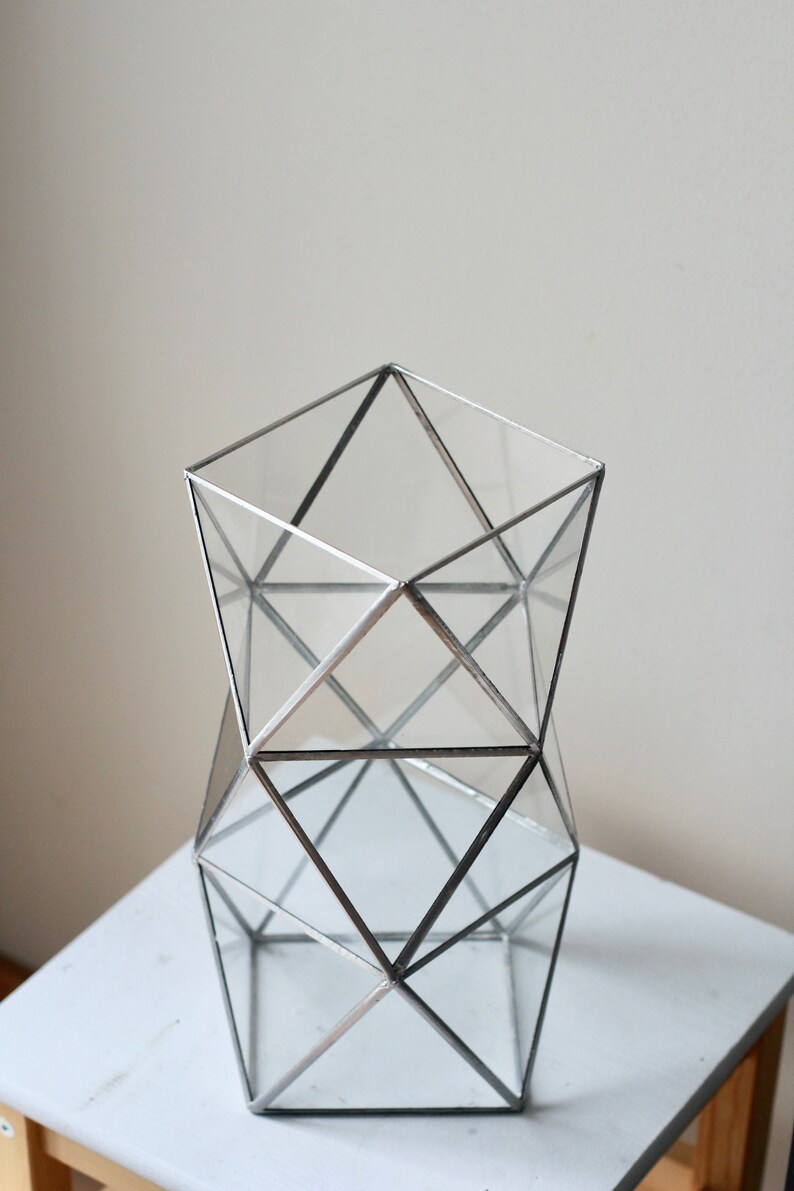 May include: A geometric glass vase with a silver metal frame. The vase is made of clear glass and has a square base. The frame is made of silver metal and has a geometric design.
