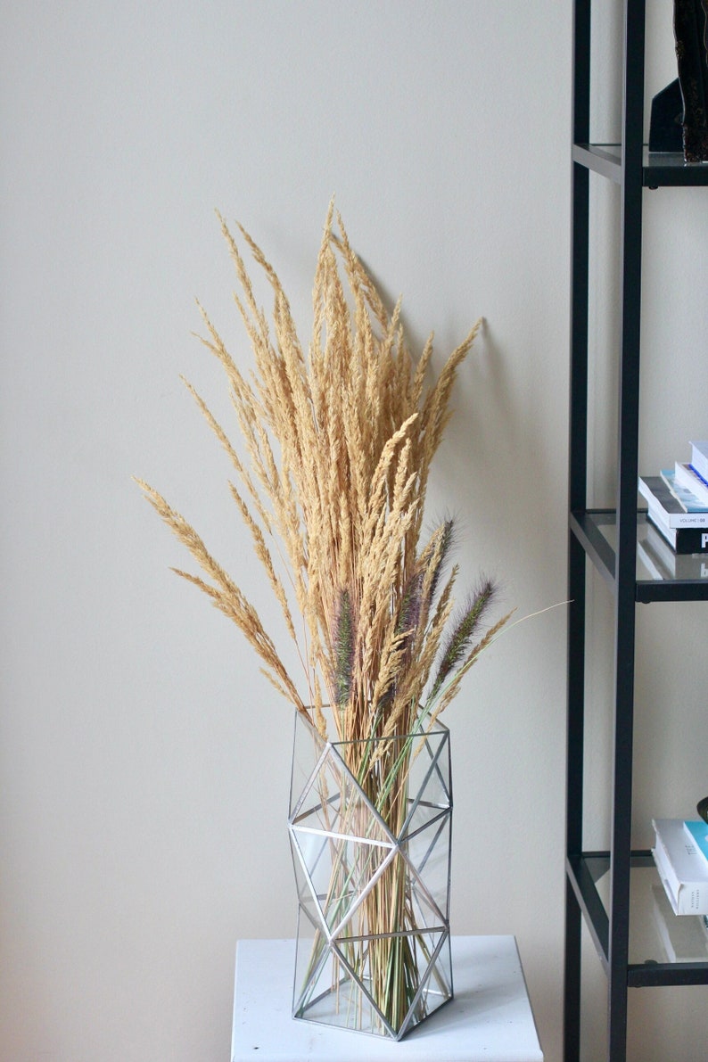 May include: A geometric glass vase with a silver frame filled with dried pampas grass and purple dried flowers.