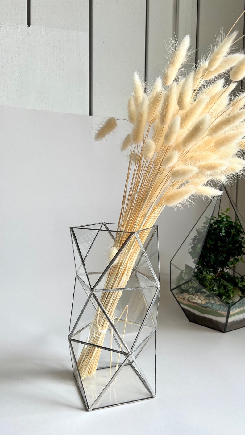 May include: A clear glass geometric vase with a silver metal frame holds a bouquet of dried white pampas grass.