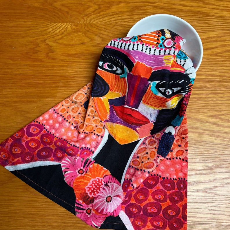 Pu&ograve; includere: Un asciugamano da cucina colorato con un design di viso stilizzato in tonalit&agrave; di rosa, arancio, rosso e blu. Il viso &egrave; circondato da un motivo di cerchi rosa e arancioni.