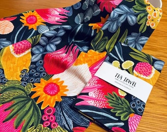 Könnte beinhalten: Ein schwarzes Geschirrtuch mit einem bunten Blumenmuster mit rosa, gelben und grünen Blumen. Das Geschirrtuch ist gefaltet und hat ein weißes Etikett mit der Aufschrift "TEA TOWEL 100% COTTON TEA TOWEL DESIGNED IN AUSTRALIA".
