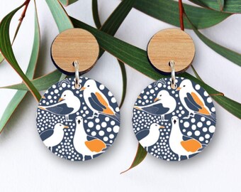 Op de afbeelding: Een paar houten oorbellen met een blauw en wit patroon van meeuwen op een witte achtergrond met witte stippen. De oorbellen zijn rond en hebben een houten bovenkant.