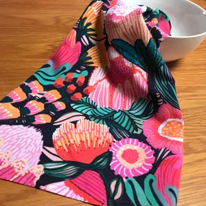 Può includere: Un tovagliolo di stoffa a fantasia floreale colorata con sfondo scuro. Il tovagliolo presenta grandi fiori stilizzati nelle tonalità del rosa, dell'arancione e del verde. Il tovagliolo è piegato e appoggiato su una superficie di legno.
