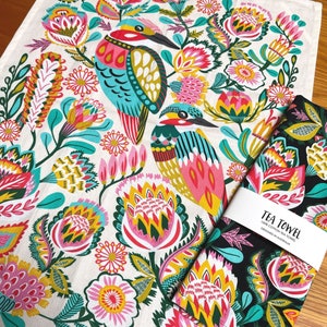 Può includere: Un asciugamano da tè bianco con un design floreale e di uccelli colorato. Il design presenta colori turchese, rosa, giallo e verde. L'asciugamano da tè ha il testo "TEA TOWEL 100% COTTON TEA TOWEL DESIGNED IN AUSTRALIA" stampato su di esso.