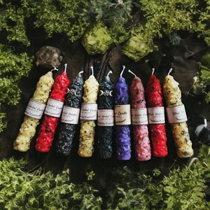 Peut inclure: Une collection de bougies aux herbes de différentes couleurs, dont le jaune, le rouge, le noir, le bleu et le violet. Chaque bougie a une texture unique et porte une étiquette "Herbal Candles". Les bougies sont disposées sur de la mousse verte.