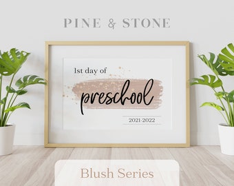 May include: A printable sign with a blush pink watercolor background and gold glitter accents. The text reads "1st day of preschool 2021-2022". The sign is framed in a light wood frame and is sitting on a wooden floor.