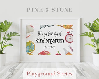 May include: A white framed print with a watercolor illustration of school supplies, including a globe, books, pencils, and an apple. The text "It's my first day of Kindergarten 2021-2022" is printed on the print. The print is part of the "Playground Series".
