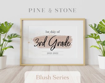 Op de afbeelding: Een printbaar bord met een roze aquarel achtergrond en gouden glitter accenten. De tekst luidt "1e dag van de 3e klas 2021-2022". Het bord is ingelijst in een licht houten lijst en wordt weergegeven op een witte muur.