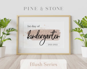 May include: A printable art print with a blush pink watercolor background and gold glitter accents. The text reads "1st day of kindergarten 2021-2022". The print is framed in a light wood frame and is displayed on a white wall with two green plants.